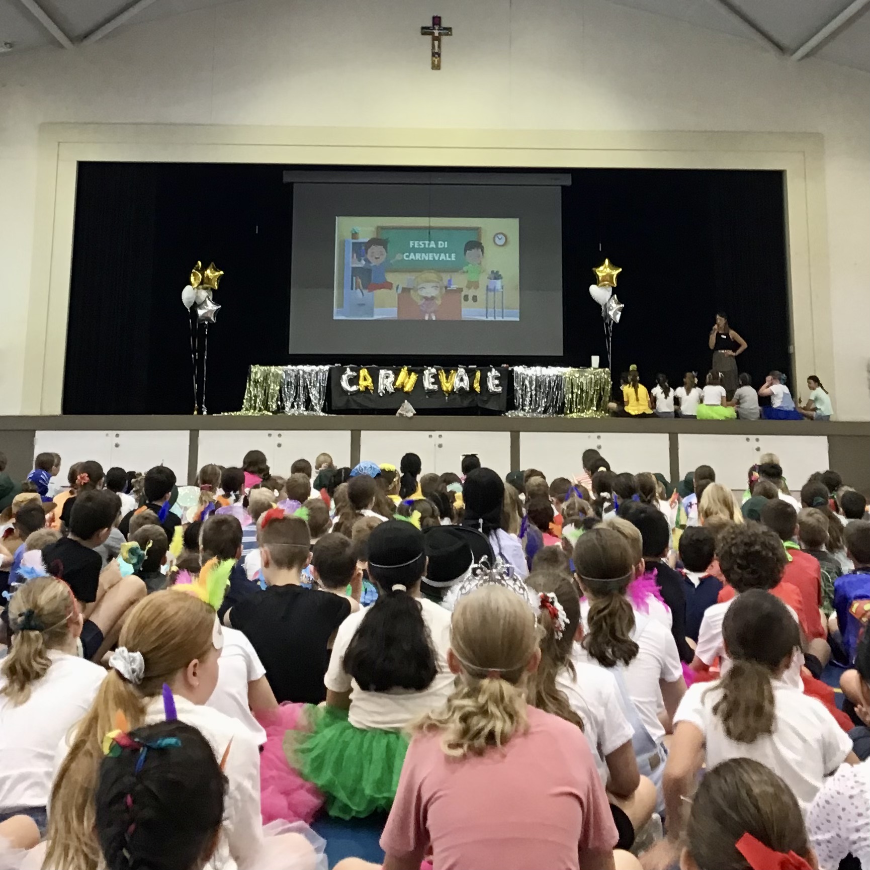 Students seated in a school hall facing a stage decorated for Carnival, with a presentation displayed on a large screen.