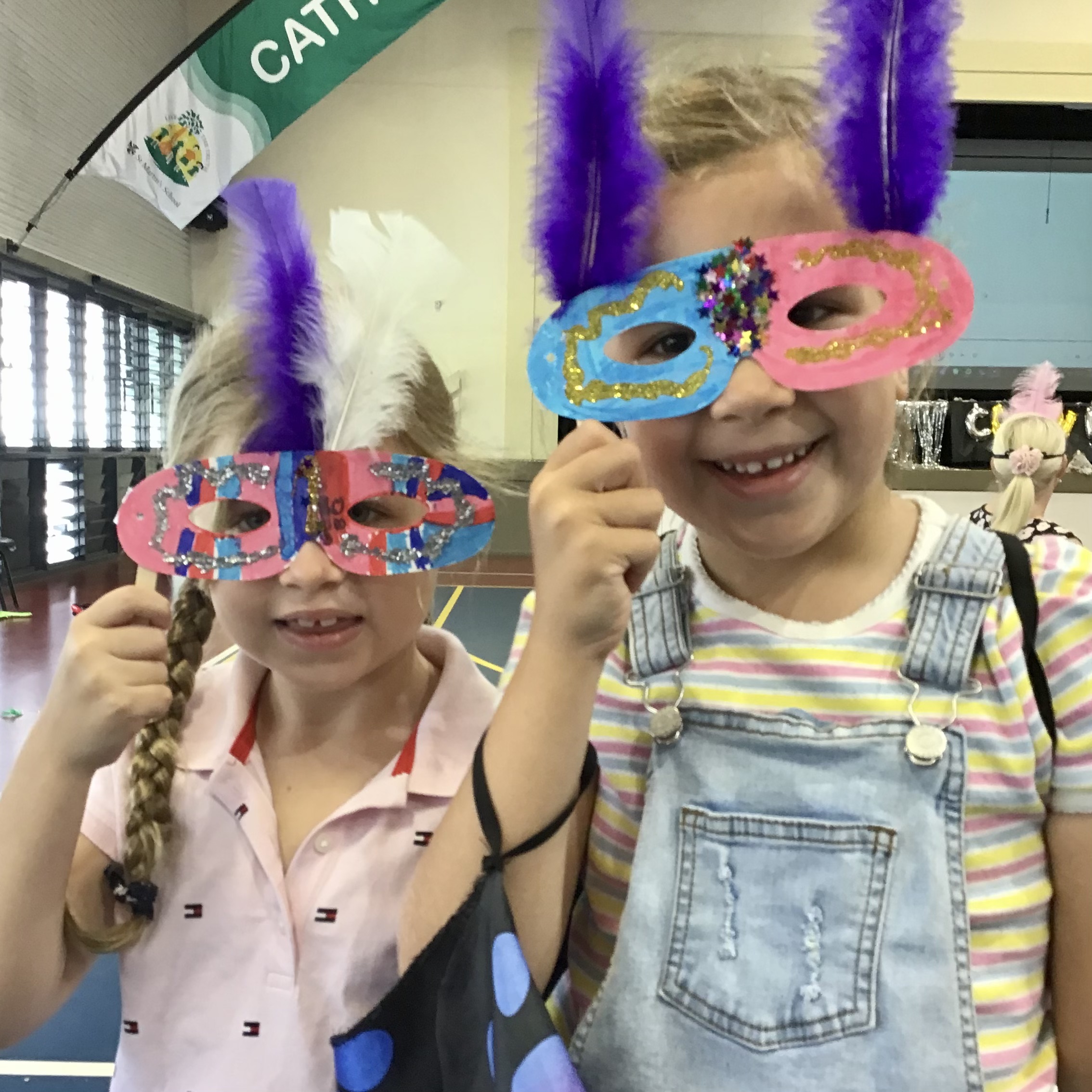 Two students standing in a school hall wearing colourful decorative masks with feathers.