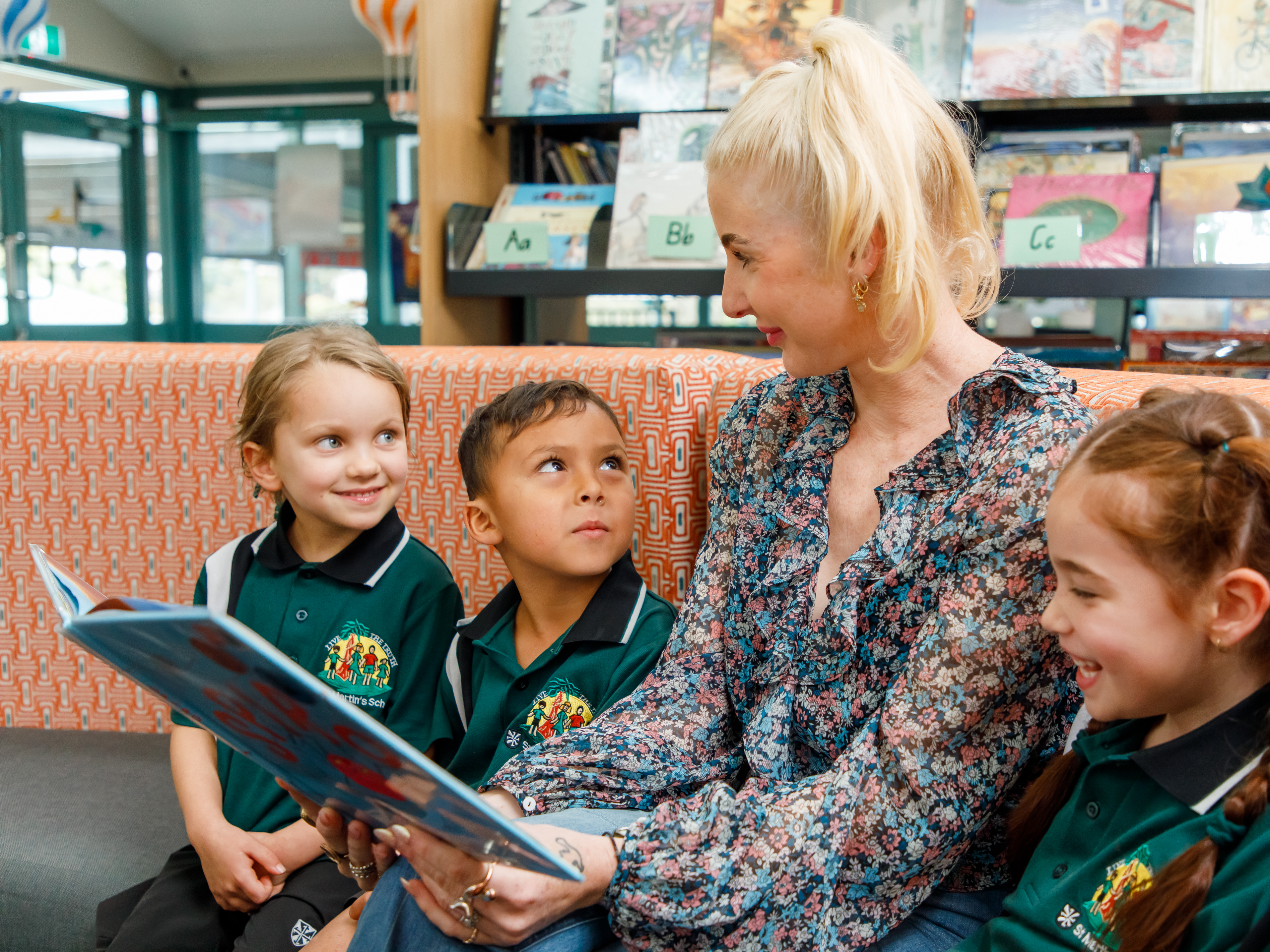 A teacher is sitting with three young students on a couch in the library. She has a book open and is reading. 
