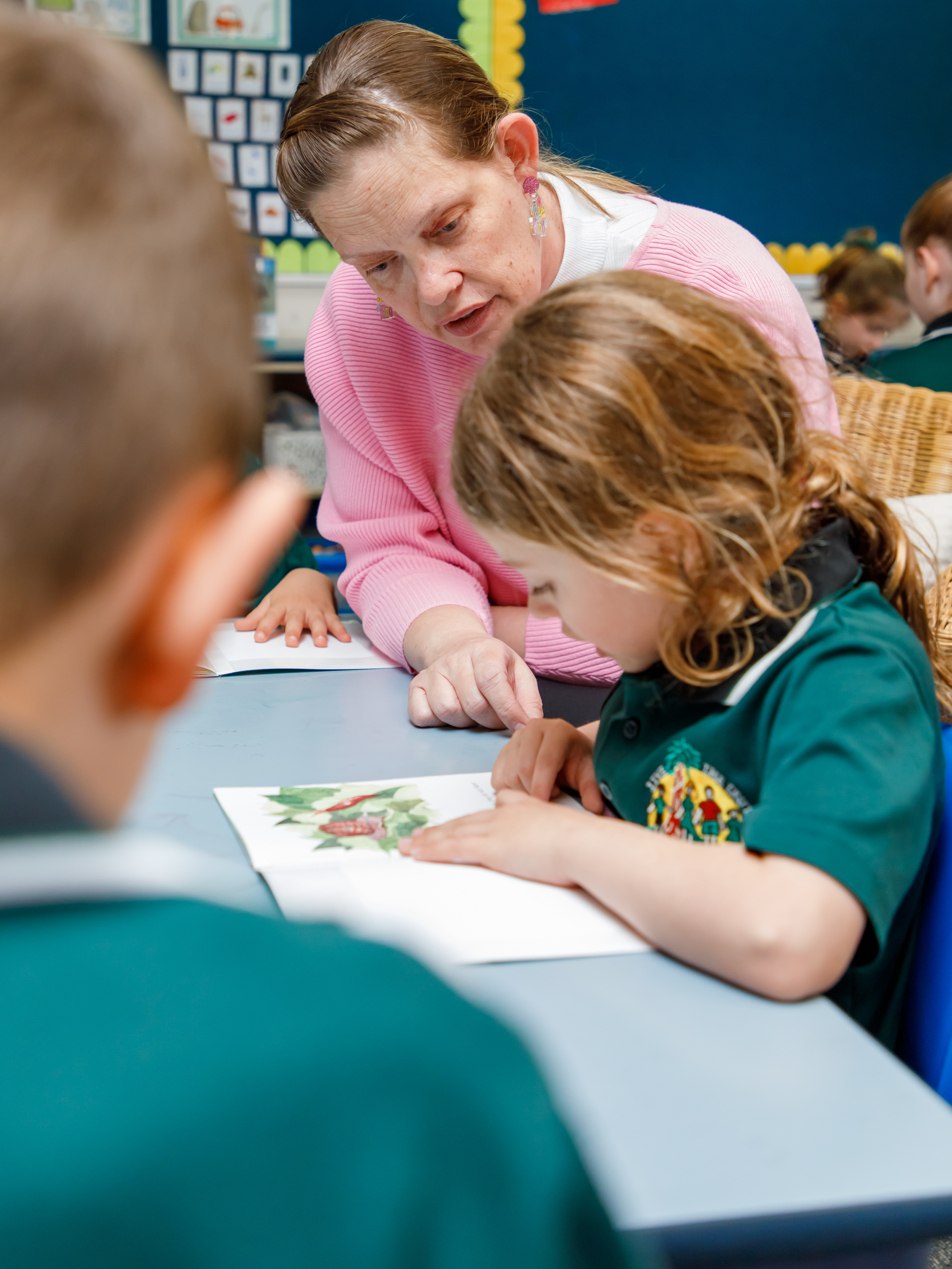 A primary aged student is sitting at a desk and reading. A teacher is standing next to her to assist.