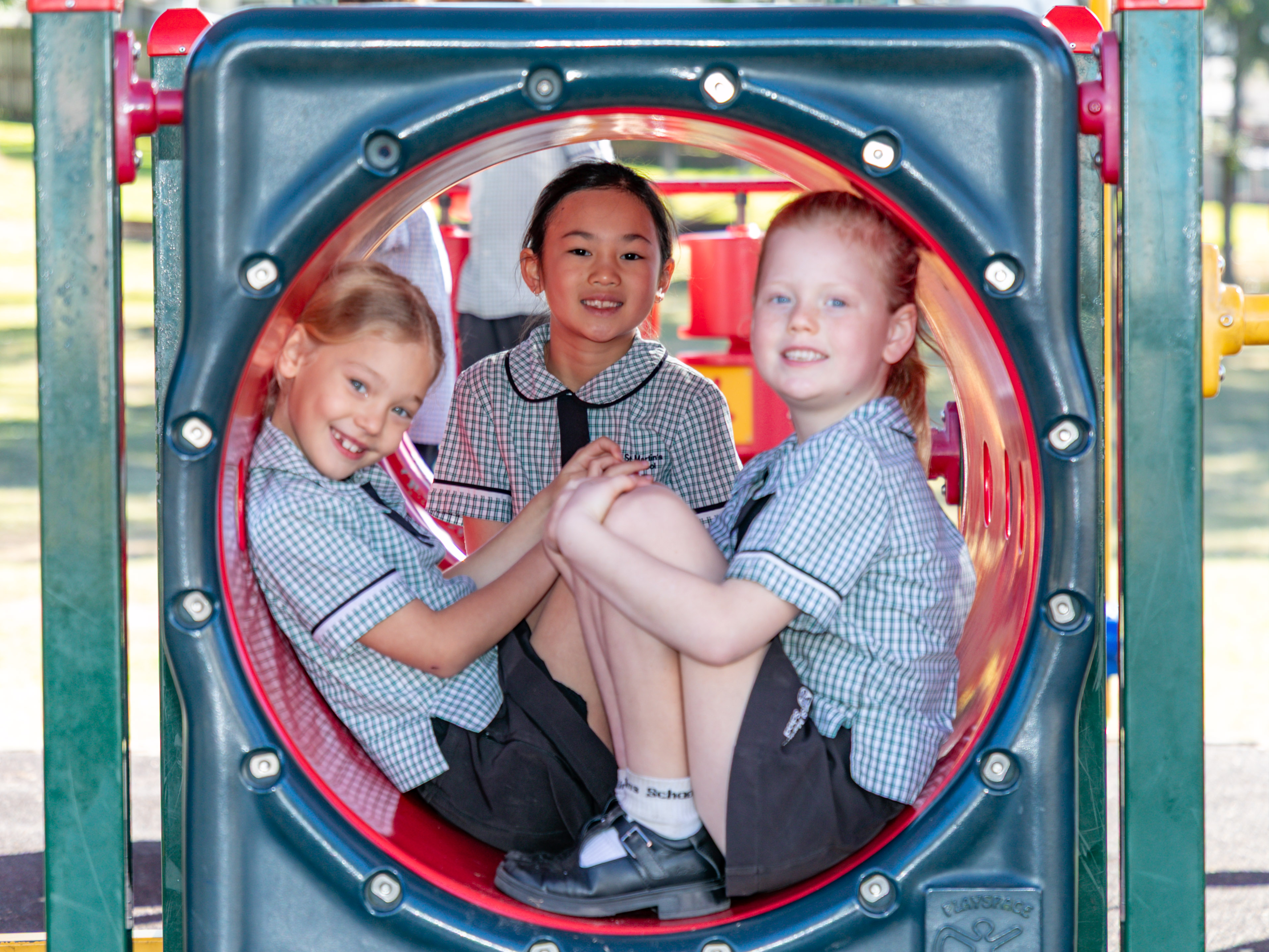 Two girls are sitting in a playground, smiling and facing towards the camera.
