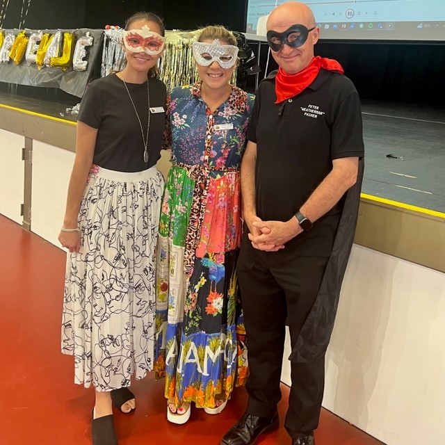 Three adults standing on a stage wearing decorative masks and costumes, with a decorated backdrop and projection screen behind them.