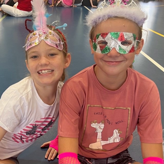 Two students seated on a gym floor wearing decorative masks and headbands, with other students seated in the background.